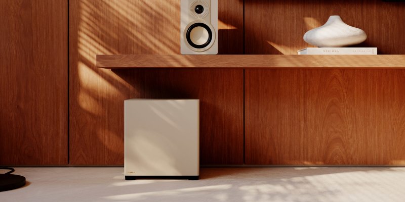 DALI BASIK SUB in Caramel White on the floor beneath a shelf-mounted speaker