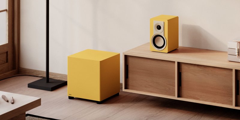 DALI BASIK SUB in Golden Yellow in a living room beside a matching standmount speaker